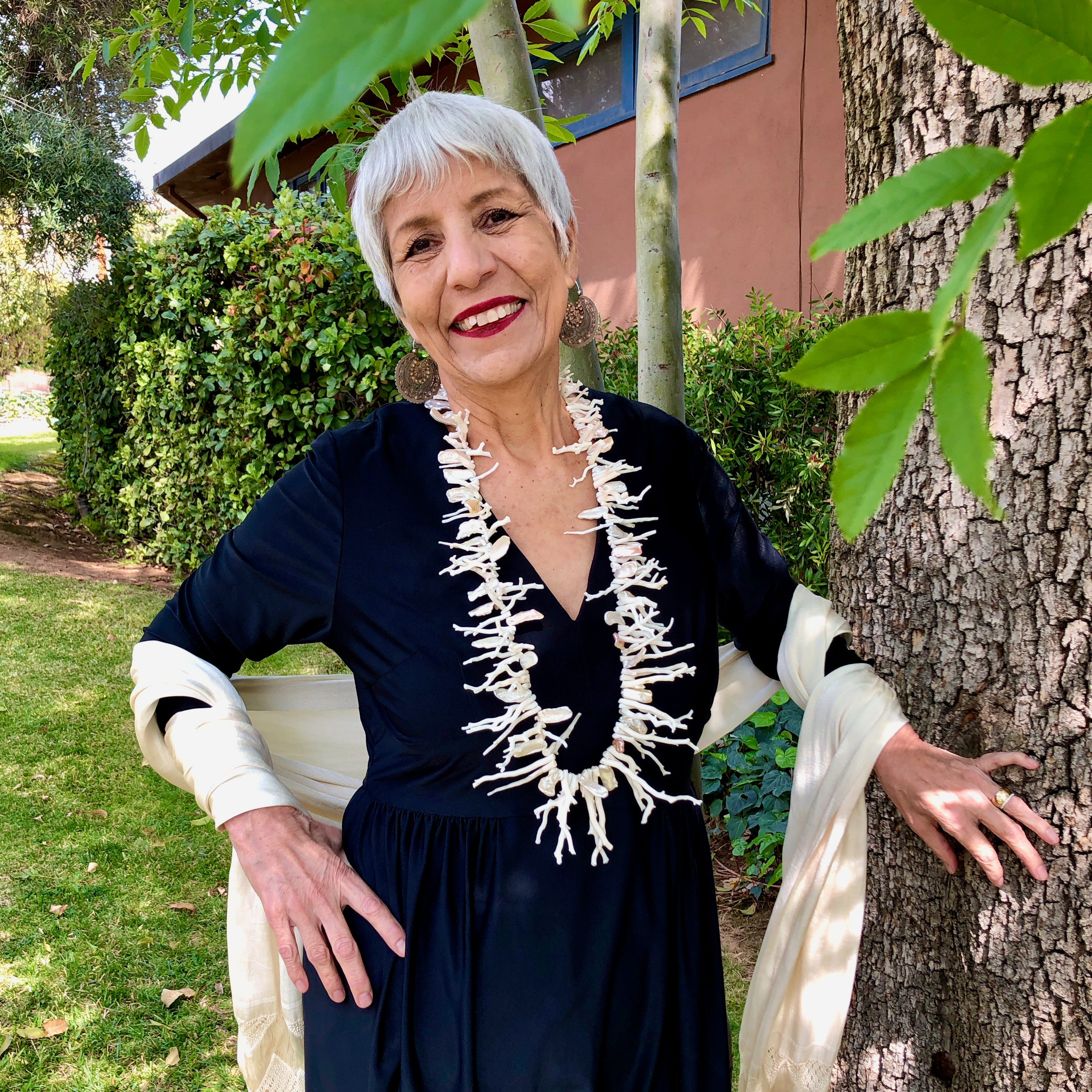 The smiling owner stands in front of green foliage with silver white hair and red lipstick wears a vintage, black, long sleeve, v-neck maxi dress and a cream colored, hand woven reboso, along with the White Branch Coral, Keshi Pearls, Sterling Silver Clasp with Zircons and Pearl Necklace, showing off the 36 inch necklace that sits at her belly at its longest point.