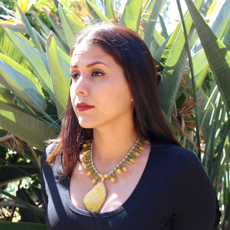 A brunette model with tan skin, red lipstick, black eyeliner, dark brown eyes in a black long sleeve, scoop neck top wearing the Butterscotch Baltic Amber Pendant with Faceted Peridot Necklace, sitting outside in front of a lush bird of paradise plant, like a Frida Kahlo portrait. The necklace sits right in the center of her chest, just below the collar bone with the large 3 inch pendant falling between her cleavage.
