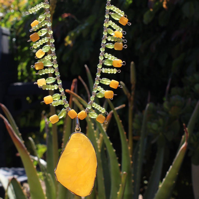 An eye level shot of the Butterscotch Baltic Amber Pendant with Faceted Peridot Necklace hanging in front of various outdoor patio plants, showing off the slight transparency of the pendant, which radiates a bright yellow glow as it soaks up the sunlight.