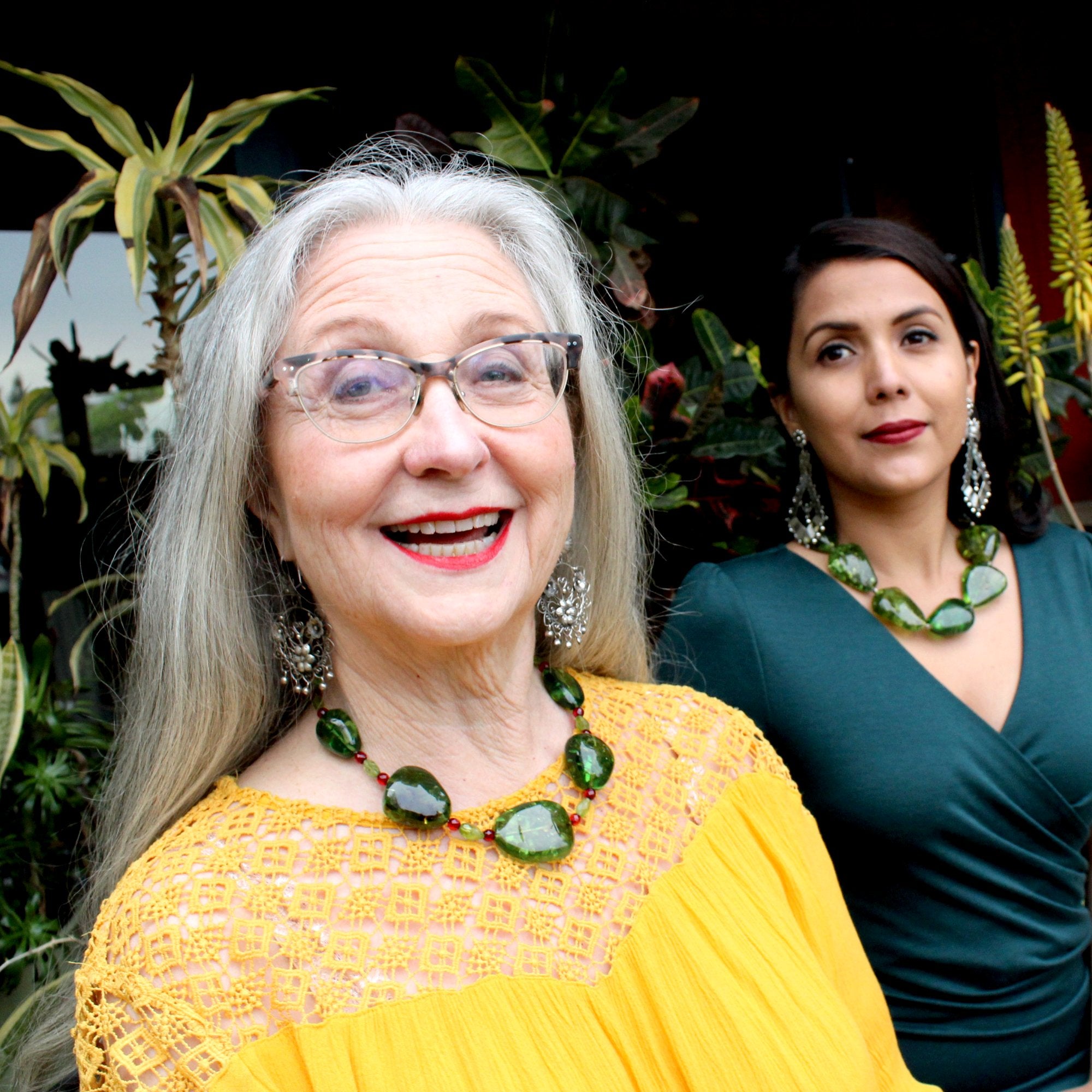 Two models are shown wearing two different variations of my Caribbean Green Amber necklaces, the one in this listing is worn by a smiling, silver haired, older model with white skin, red lipstick, black eyeliner, blue eyes, in a yellow crochet crew neck top. The necklace falls over her top, right in the center of her chest, just below the collar bone. They both sit in front of various succulents and other foliage.