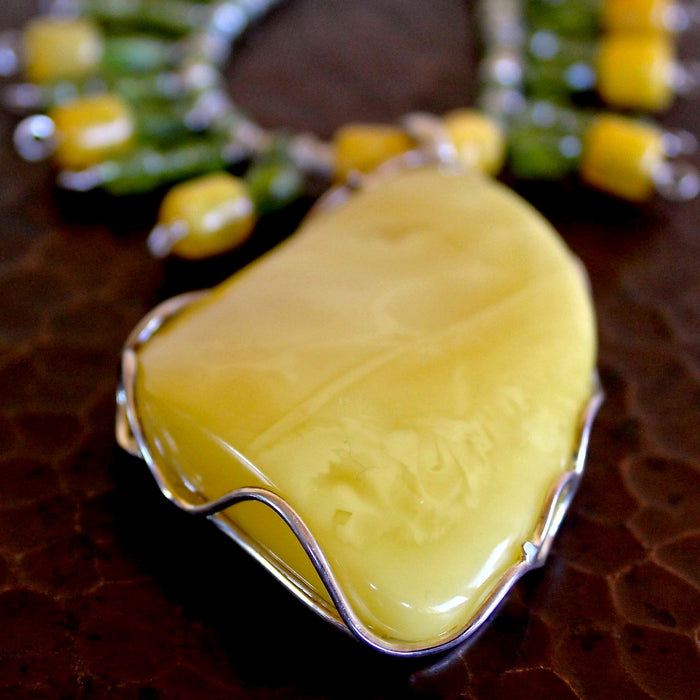 Close up shot of the Butterscotch Baltic Amber Pendant with Faceted Peridot Necklace, showing off the incredible creamy yellow amber pendant in great detail, including the sterling silver wire work along the edges. The shot is taken at a 45 degree angle overhead, with the necklace sitting on a hand hammered copper table with the pendant in the foreground, and the rest of the necklace out of focus, receding into the background.