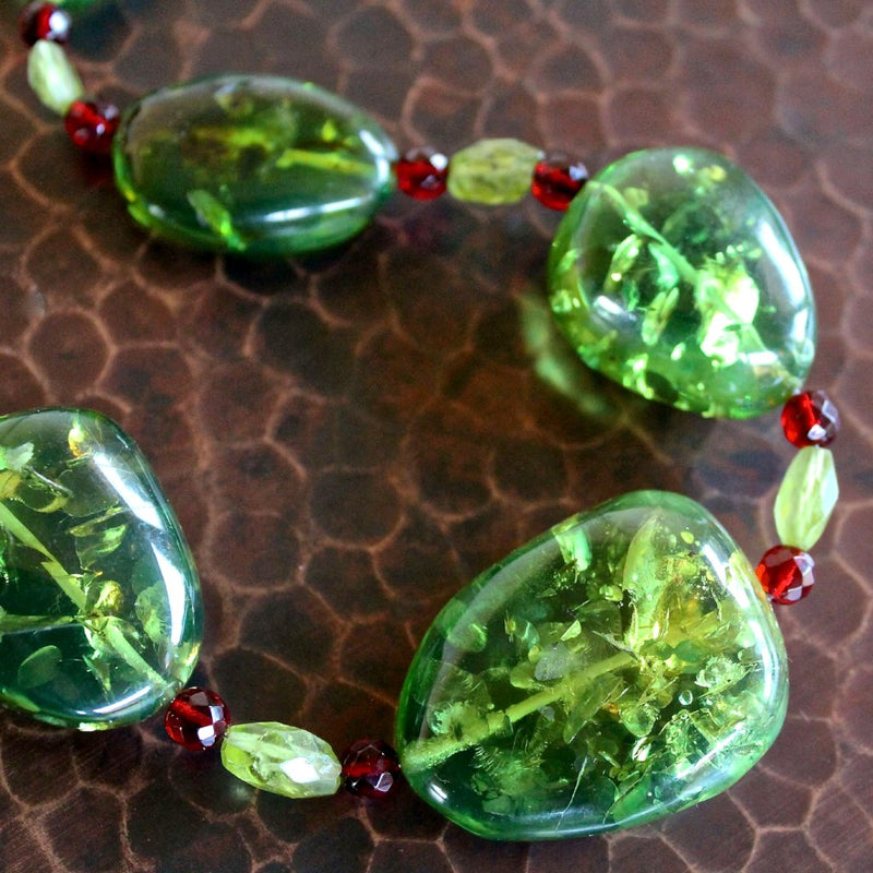 An overhead, in-focus, detail shot of the Caribbean Green Amber, Red Baltic Amber and Faceted Peridot Bead Necklace. Each piece of amber is a variation of rich, kelly green and lighter lime green, translucent, and uniquely oblong shaped with lots of detail and texture inside each smooth bead. The small, faceted red baltic amber and pale green peridot beads sparkle in the light. The necklace sits on a dark brown hand hammered copper table.
