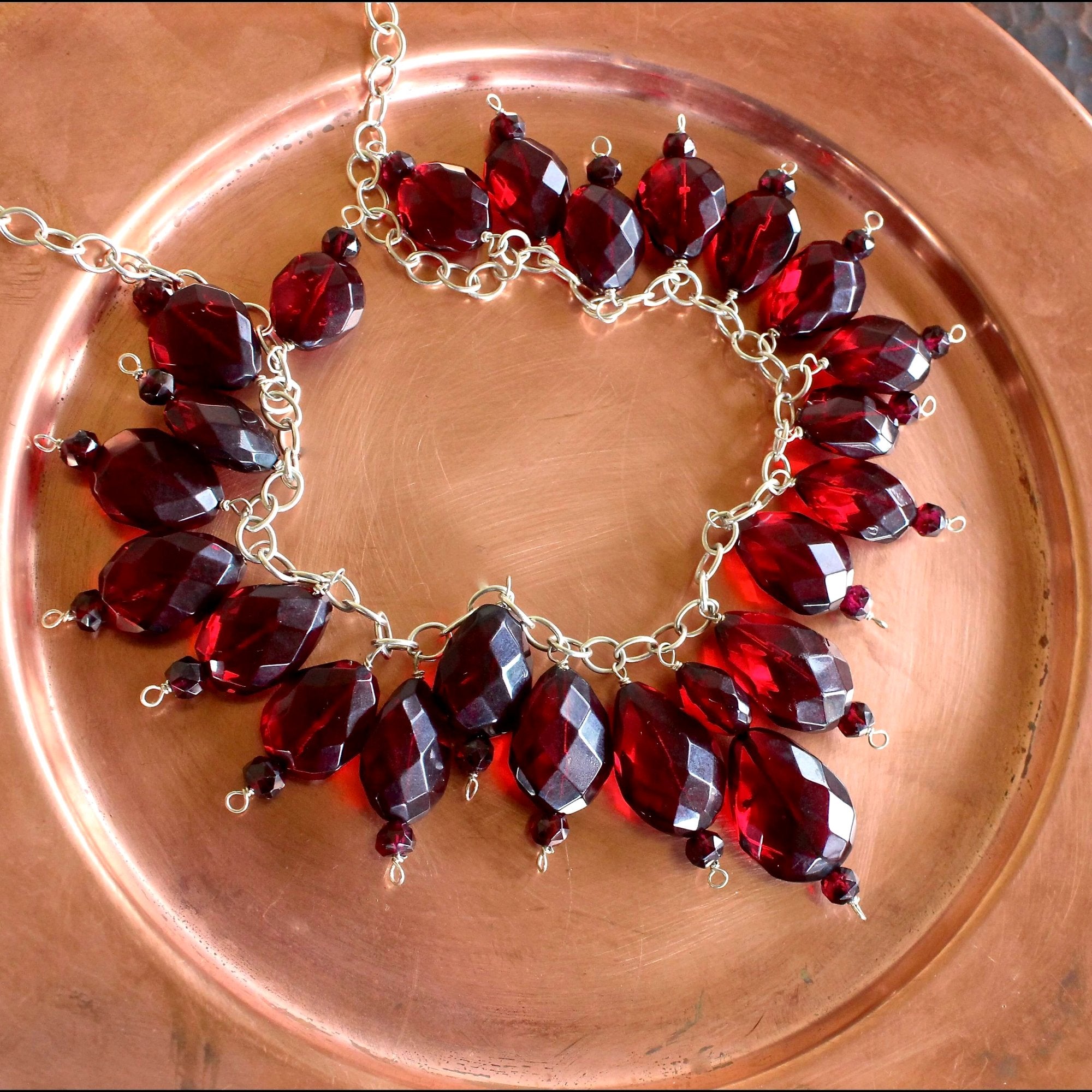 An overhead, in-focus, close-up shot of the Faceted Red Baltic Amber, Red Garnet, Sterling Silver and Hill Tribe Silver Beaded Necklace, showing the luminescence and vibrant red color of the amber beads in diffused natural daylight. The necklace sits on a matte copper dish.