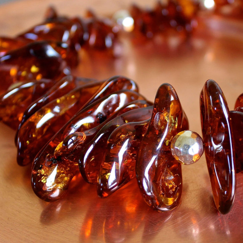Close up shot of the translucent, smooth, rich, warm, flattened, oblong shaped whiskey amber beads and one in-focus hand hammered pure Hill Tribe silver bead. The shot is taken at eye level with the necklace sitting on a matte copper plate that gives off a faint reflection of the necklace.