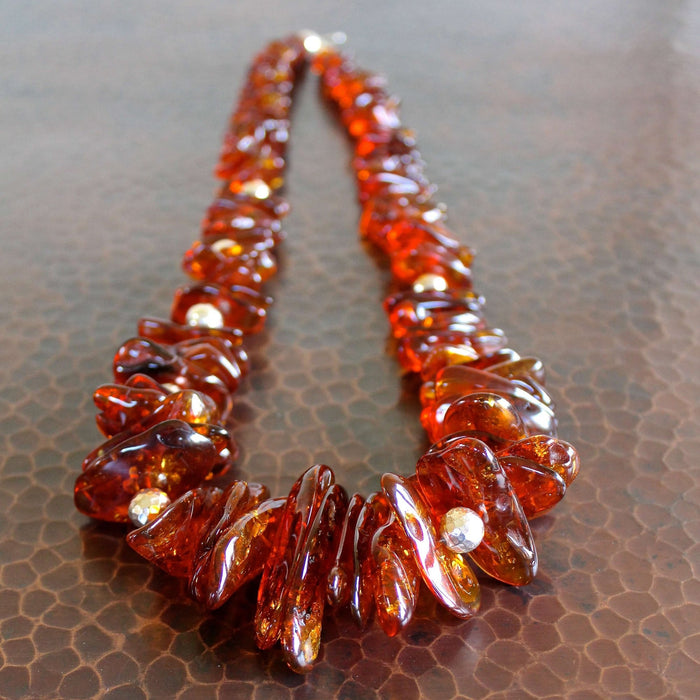  An almost eye level shot of the Whiskey Baltic Amber with Hill Tribe Silver Necklace in full, but the clasp of the necklace is out of focus in the background while the focal point of the necklace is in the foreground, laying on a hand hammered copper table.