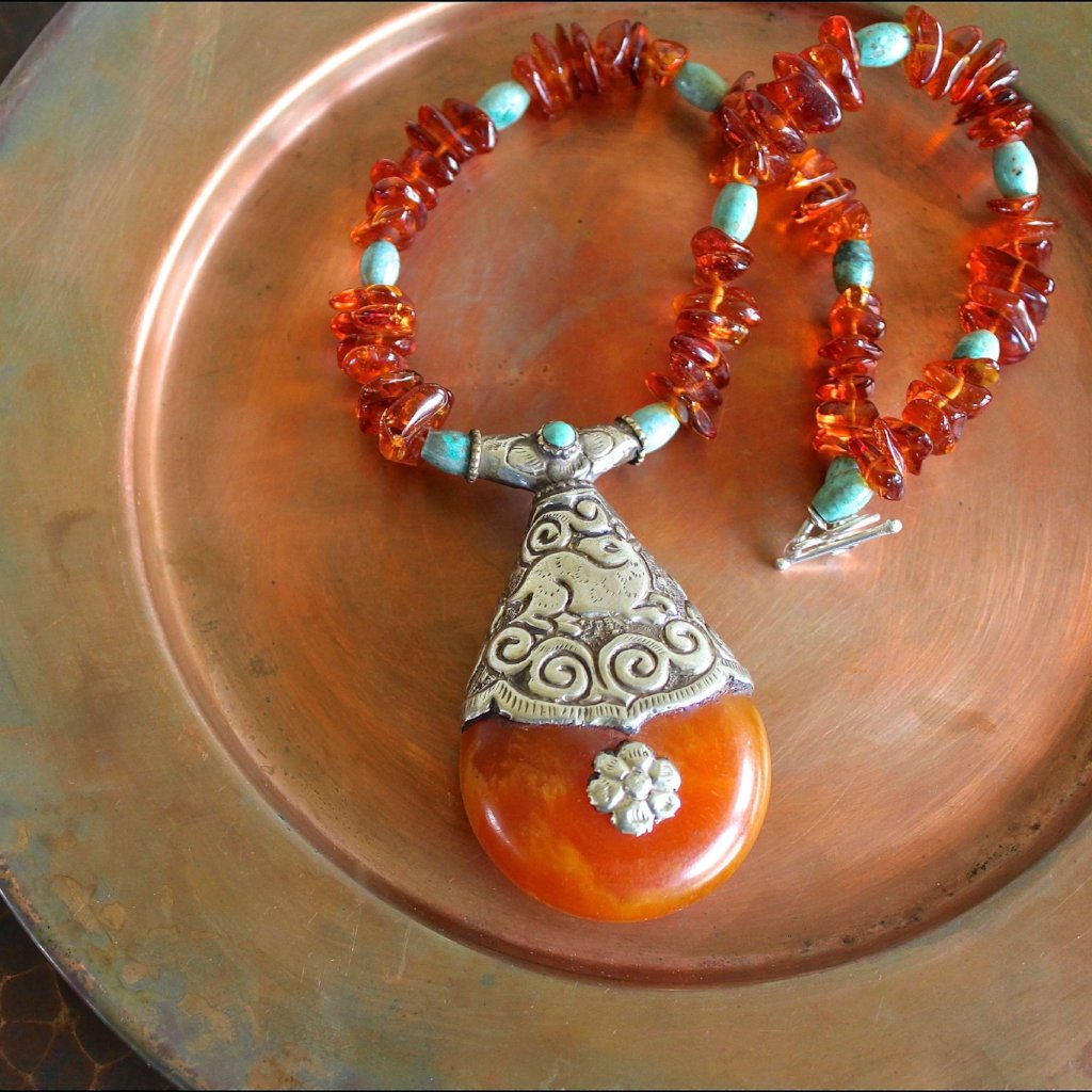 An overhead shot of the Tibetan Copal Pendant, Cognac Baltic Amber, and Turquoise Necklace, with the entire necklace in focus, laying on a matte copper plate. Featuring a Tibetan pendant of copal, silver and turquoise with cognac Baltic amber beads, green turquoise beads, sterling silver toggle clasp. The pendant showcases spirals, a flower, and the image of the doe on the silver work. 