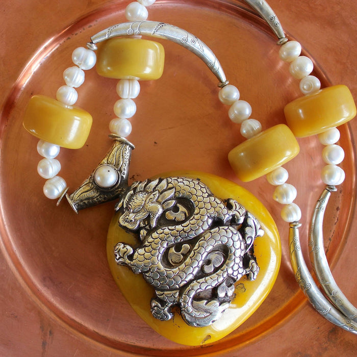 Close up shot of the Tibetan Double-Sided Yellow Copal Pendant with Carp and Dragon, Pearls, Large Copal Beads Necklace, showing off the incredible, opaque, creamy yellow copal pendant in great detail, including the intricate silver dragon design on the pendant, the light texture in the pearl beads, the four large cylinder shaped copal beads, and the curved and carved Hill Tribe pure silver spacers. The necklace is sitting folded on a matte copper plate
