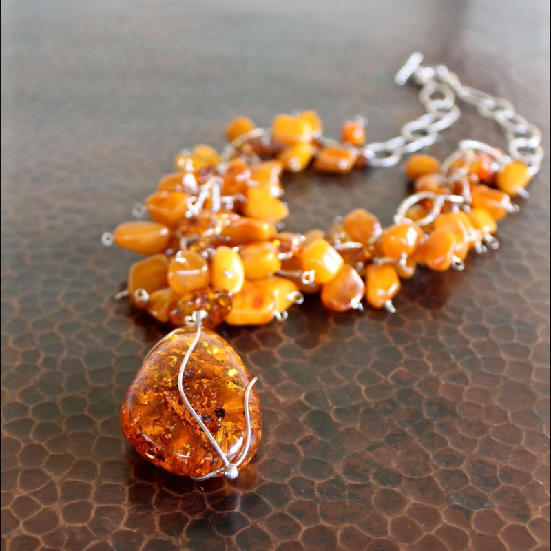 An almost eye level shot of the in-focus Cognac Baltic amber pendant (3") wrapped in sterling silver, showcasing the detail. On sterling silver chain are butterscotch Baltic amber and cognac Baltic amber beads handwired to the loops of the chain, with an out of focus sterling silver toggle clasp shown in the background.