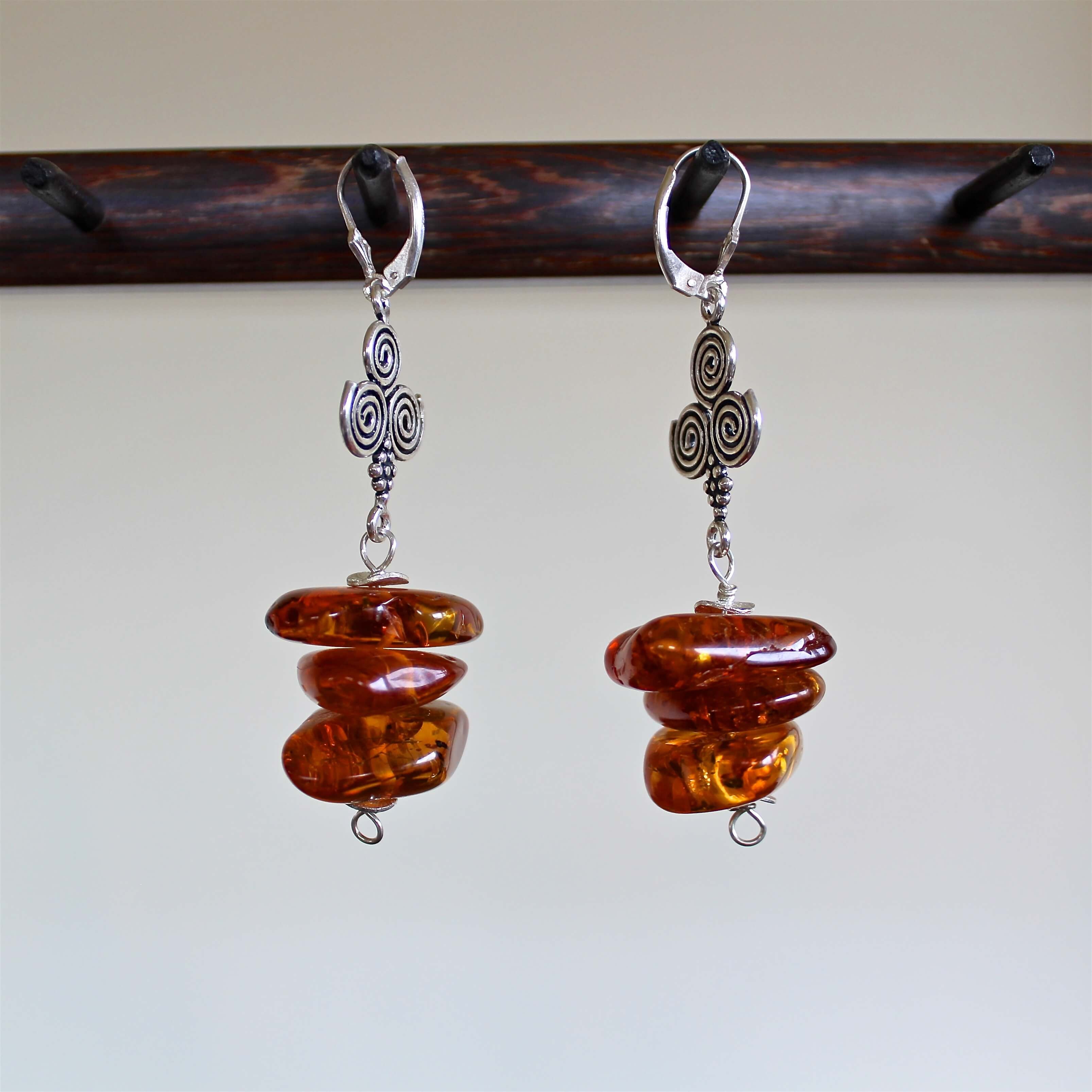 An eye level shot of the Cognac Baltic Amber and Sterling Silver Spiral Bead Earrings hanging in front of a white background showing off the various rich, reddish brown tones of these stacked, intentionally mismatched, one of a kind oblong shaped amber beads, along with the detail of the sterling silver spiral beads.