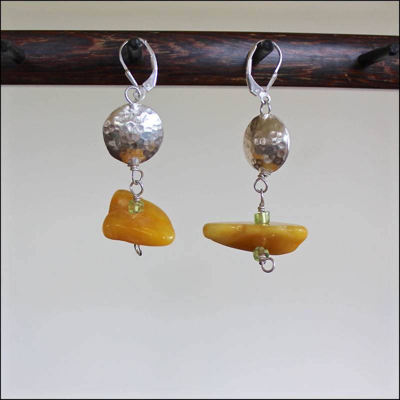 An eye level shot of the Butterscotch Baltic Amber, Peridot and Hill Tribe Silver Bead Earrings hanging in front of a white background showing off the various creamy yellow tones of these intentionally mismatched, one of a kind oblong shaped amber beads in various sizes, along with the detail of the small peridot and hand hammered Hill Tribe pure silver beads.