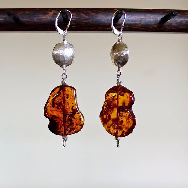 An eye level shot of the Cognac Baltic Amber with Hill Tribe Silver Bead Earrings hanging in front of a white background showing off the various golden brown tones of these mismatched, one of a kind oblong shaped amber beads, along with the detail of the hand hammered Hill Tribe pure silver beads.