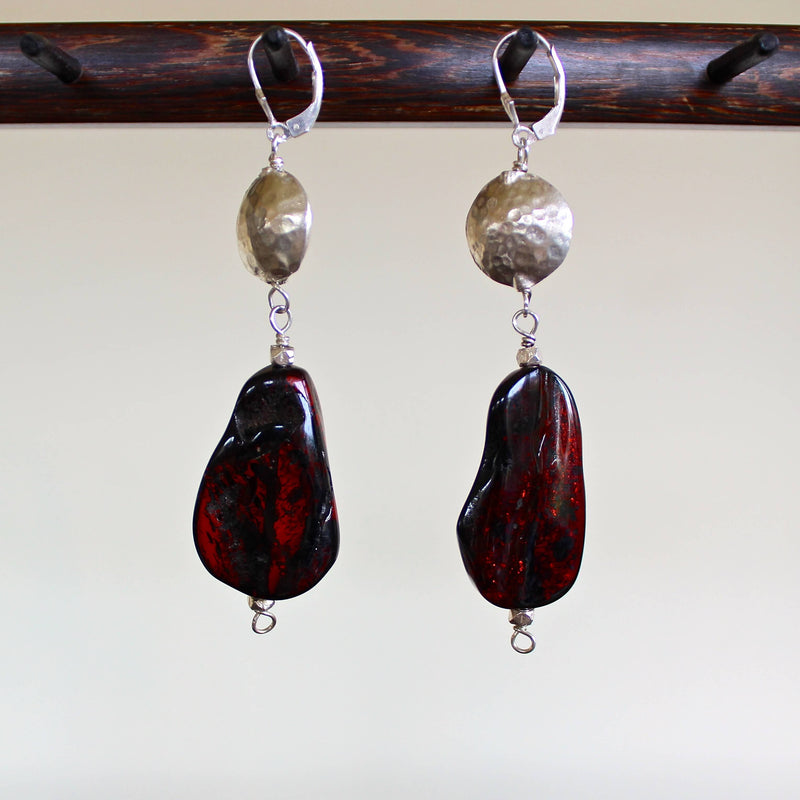 An eye level shot of the Whiskey Baltic Amber with Hill Tribe Silver Earrings hanging in front of a white background showing off the deep, rich reddish brown tones of these one of a kind oblong shaped amber beads, along with the detail of the hand hammered Hill Tribe pure silver beads.