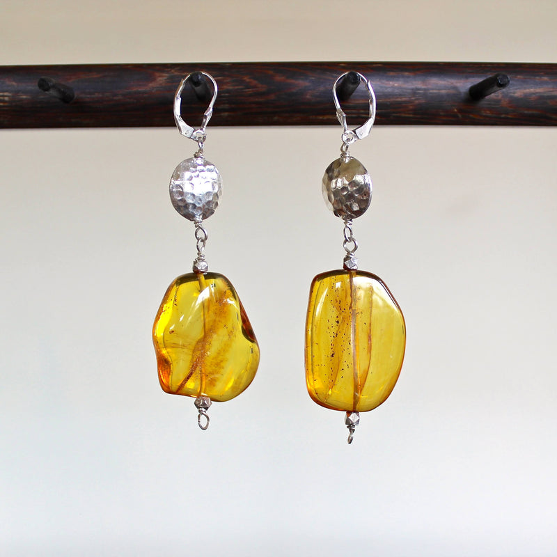 An eye level shot of the Honey Baltic Amber and Hill Tribe Silver Earrings hanging in front of a white background showing off the intricate texture and swirls inside of each translucent, vibrant, golden one of a kind amber bead, and the hand hammered detail on the Hill Tribe pure silver beads.
