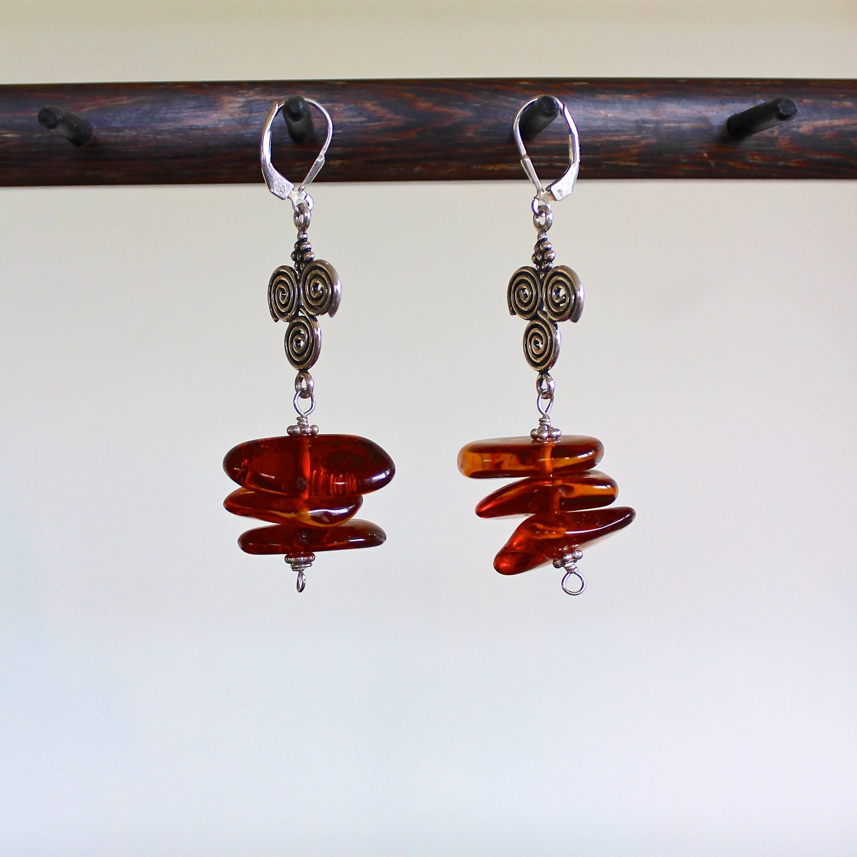 An eye level shot of a pair of Baltic Cognac Amber and Sterling Silver Spiral Pendant Earrings hanging on a dark wooden stand in front of a white wall. The earrings feature rich, reddish brown cognac amber beads, flat sterling silver charms with three spiral shapes, two on top, one on bottom, sterling silver beads with sterling silver lever back ear wires