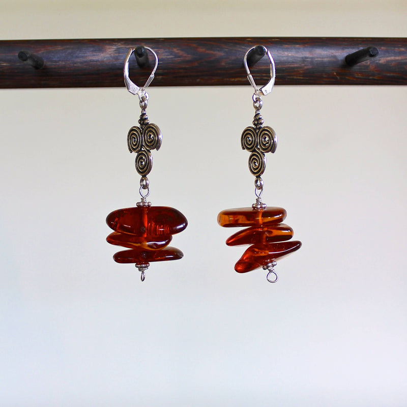 An eye level shot of a pair of Baltic Cognac Amber and Sterling Silver Spiral Pendant Earrings hanging on a dark wooden stand in front of a white wall. The earrings feature rich, reddish brown cognac amber beads, flat sterling silver charms with three spiral shapes, two on top, one on bottom, sterling silver beads with sterling silver lever back ear wires