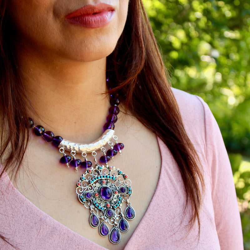 A brunette model with tan skin, berry pink lipstick, in a long sleeve, v-neck pale pink top is wearing the Matilde Poulat-Style Pendant with Hill Tribe Pure Silver and Faceted Amethyst Beaded Necklace, sitting outside with lots of greenery behind her. The necklace sits right in the center of her chest, just below the collar bone, with the stones shining in the diffused sunlight. 